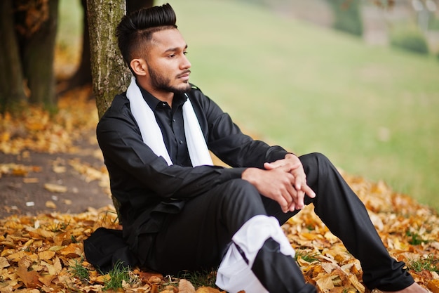 Uomo indiano alla moda in abiti tradizionali neri con sciarpa bianca in posa all'aperto contro l'albero giallo delle foglie di autunno