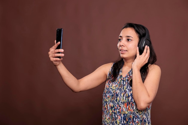 Indian woman in headphones chatting on smartphone videocall,\
talking on teleconference. person wearing wireless earphones,\
holding mobile phone, having online meeting conversation