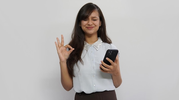 写真 インドの女性がスマートフォンを持ち、グレイの背景にウインクしながらokサインを身振りで示す