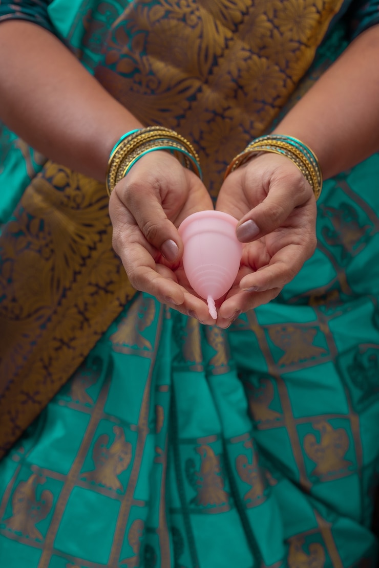 फोटो एक भारतीय महिला पुन: प्रयोज्य सिलिकॉन मेन्स्ट्रुअलकप पकड़े हुए है और विभिन्न प्रकार के फोल्डिंग दिखा रही है।