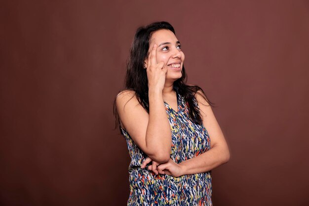 Indian woman laughing, holding finger on face, looking upwards
portrait. pensive cheerful model standing with carefree facial
expression, happy lady thinking, studio medium shot