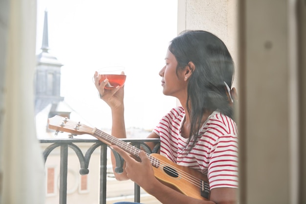 写真 ティーカップを飲みながら窓辺でウクレレを演奏するインドの女性