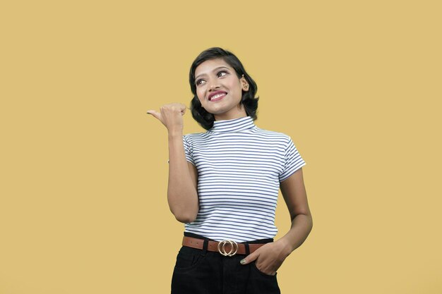 Indian woman pointing thumb at copy space over yellow\
background looking sideways pointing finger