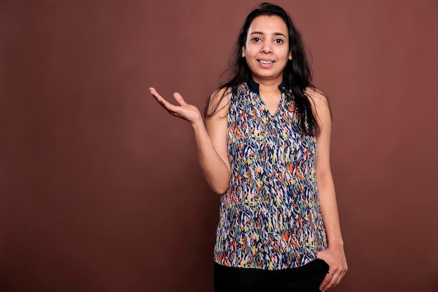 Indian woman smiling, advertising product with raised arm. Lady standing, looking at camera, showing promotion gesture portrait, front view studio medium shot on brown background