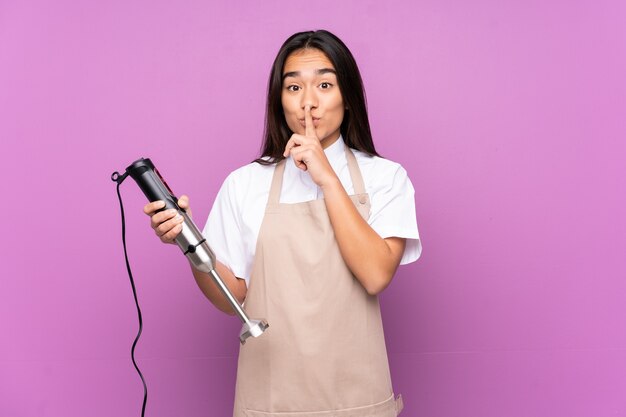 Indian woman using hand blender isolated on purple background doing silence gesture