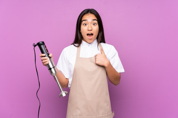 Indian woman using hand blender isolated on purple background with surprise facial expression