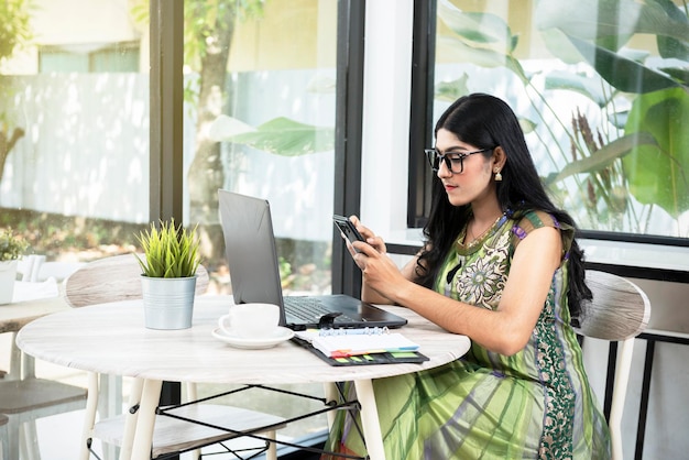 Indian woman with eyeglass using a mobile phone with a laptop and a notebook with a cup of coffee on the table