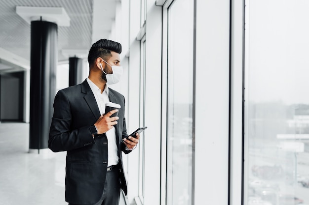 An Indian young businessman in a protective mask, in an office near a panoramic window, solves work problems with a phone in his hands and with coffee.