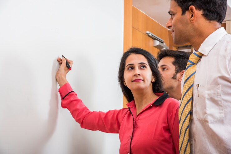 फोटो indian young businesswoman writing over white board with marker pen and other businesspeople looking