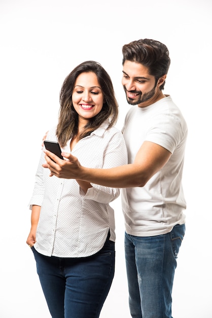 Indian young couple using smartphone or mobile handset, standing isolated over white background or against red brick background