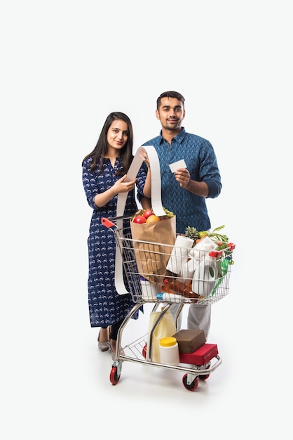 ショッピングカートまたは食料品、野菜、果物でいっぱいのトロリーを持つインドの若いカップル。白い壁の上の孤立した全身写真