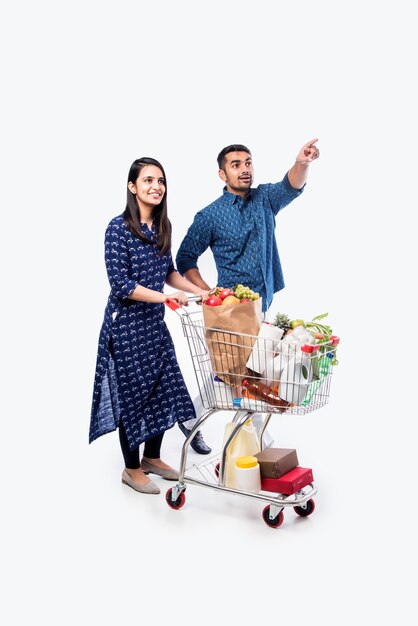 Indian young couple with shopping cart or trolly full of grocery, vegetables and fruits.  Isolated Full length photo over white wall