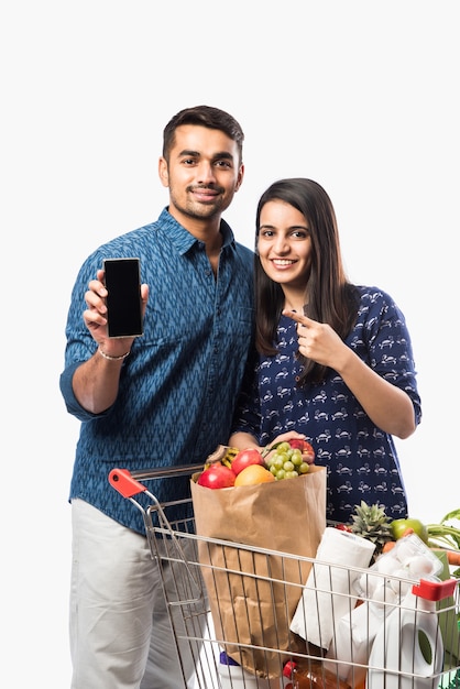 Indian giovane coppia con carrello o carrello pieno di generi alimentari, frutta e verdura. isolato foto a figura intera su muro bianco