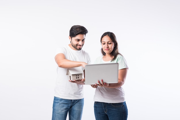 Indian young family couple choosing new home online, search real estate to buy or rent, house for sale on screen of computer, standing against white background