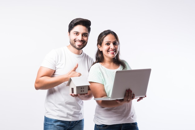 Indian young family couple choosing new home online, search real estate to buy or rent, house for sale on screen of computer, standing against white background