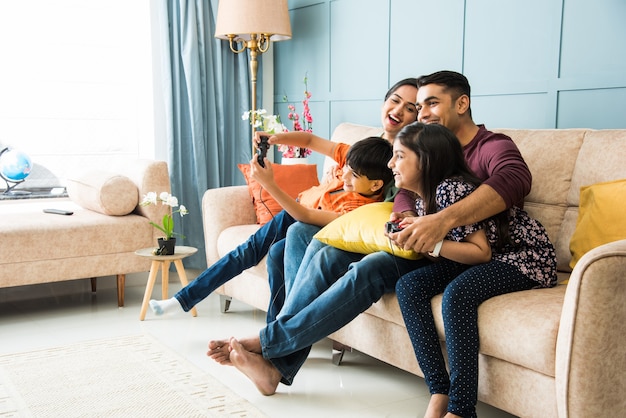 Indian young family of four playing video game using controller or joystick while sitting on sofa