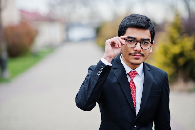 Indian young man at glasses, wear on suit with red tie posed outdoor.