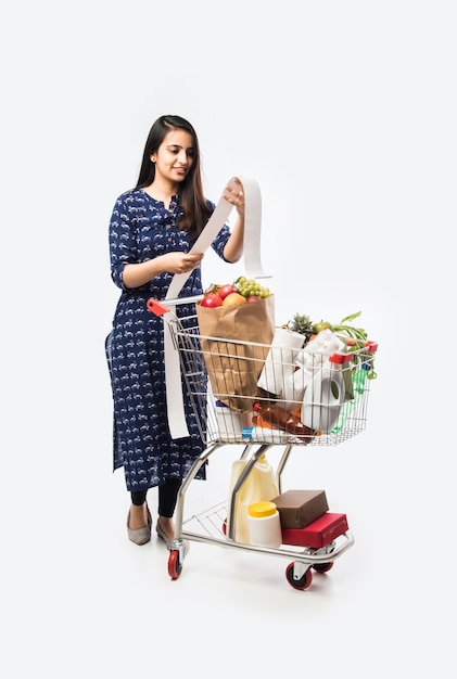 ショッピングカートまたは食料品、野菜、果物でいっぱいのトロリーを持つインドの若い女性。白い壁の上の孤立した全身写真