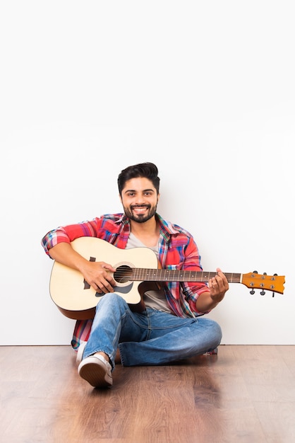 Indiase Aziatische bebaarde jonge man die akoestische gitaar speelt terwijl hij geïsoleerd zit over houten vloer tegen witte muur