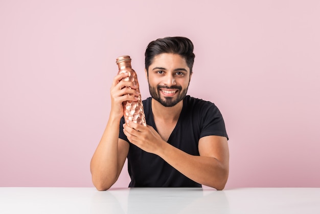 Indiase Aziatische bebaarde jonge man die een koperen waterfles vasthoudt of presenteert tegen een roze achtergrond terwijl hij aan tafel zit