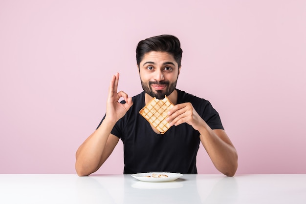 Indiase Aziatische jonge bebaarde man eet broodje brood terwijl hij in de keuken of eettafel zit. Tonen of presenteren