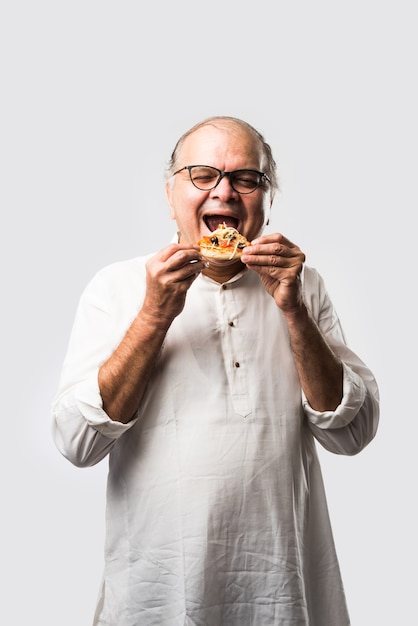 Indiase Aziatische senior of oude man die pizza eet terwijl hij geïsoleerd staat tegen een witte muur