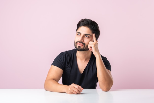 Indiase bebaarde jonge man denken zittend geïsoleerd tegen roze achtergrond aan tafel of bureau