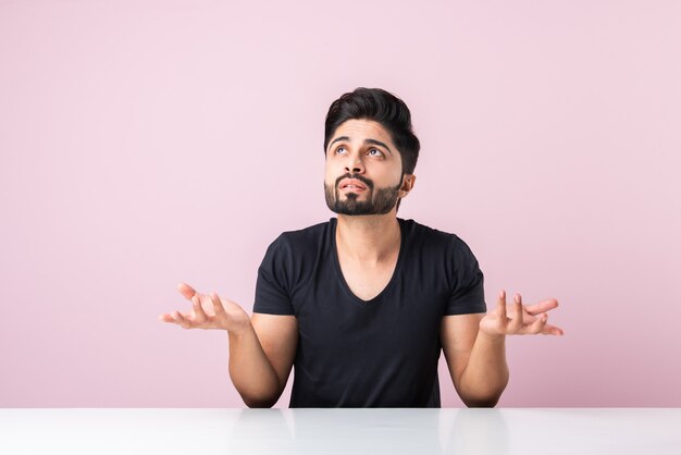 Indiase bebaarde jonge man denken zittend geïsoleerd tegen roze achtergrond aan tafel of bureau
