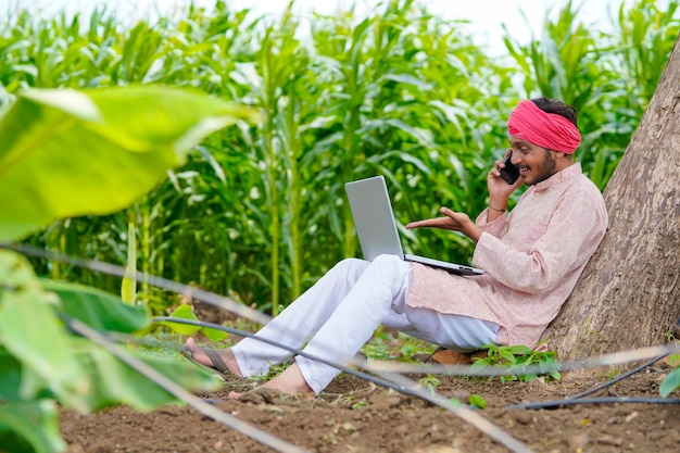 Indiase boer met behulp van laptop en praten op smartphone op landbouwveld.