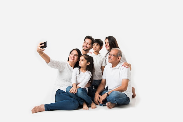 Indiase familie die selfie-foto maakt met smartphone terwijl ze op een witte achtergrond zit, omvat 3 generaties. selectieve focus