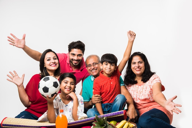 Indiase familie geniet van picknick binnenshuis - Multi generatie Aziatische familie zit op een witte achtergrond met fruitmand, mat, voetbal en drankjes. selectieve focus