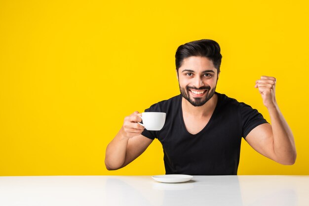 Indiase jonge man die een theekopje vasthoudt, drinkt en geniet van de aromakoffie. Aziatische man zit aan tafel tegen gele achtergrond met cup