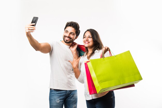 Indiase jonge paar boodschappentassen en smartphone of mobiel, op selfie klikken of winkel lokaliseren, staande geïsoleerd op witte achtergrond. selectieve focus