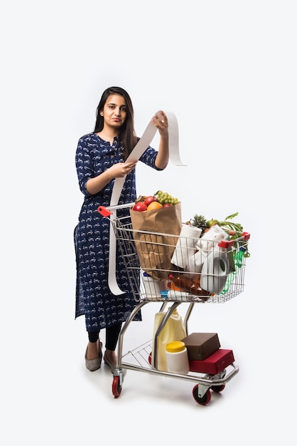 Indiase jonge vrouw met winkelwagentje of karretje vol kruidenierswaren, groenten en fruit. Geïsoleerde foto van volledige lengte over witte muur