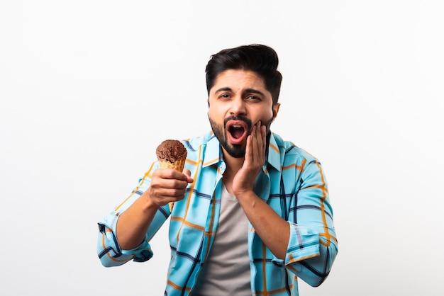 Indiase jongeman die ijs eet in kegel en tandpijn heeft vanwege gaatjes, zittend aan tafel tegen gele of witte achtergrond