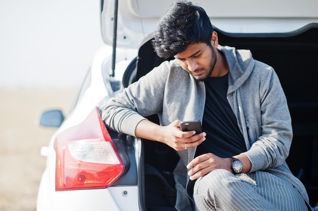 Indiase man in vrijetijdskleding poseerde voor witte auto en keek naar zijn mobiele telefoon