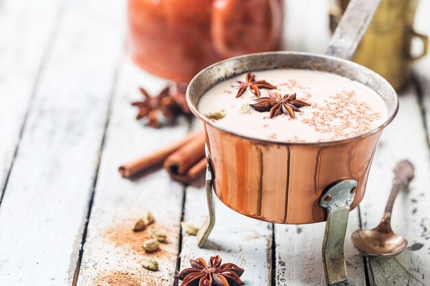 Indiase masala chai thee, gekruide thee met melk in een koperen pot op een witte houten ondergrond