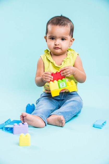 Indiase of Aziatische peuter of baby of baby die met speelgoed of blokken speelt terwijl hij ligt of zit geïsoleerd op een heldere of kleurrijke achtergrond