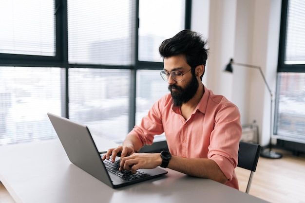 Indiase ontwerper man in stijlvolle bril werken op laptop zitten aan bureau in lichte coworking kantoorruimte door raam Bebaarde zakenman dragen casual kleding kijken naar computerscherm