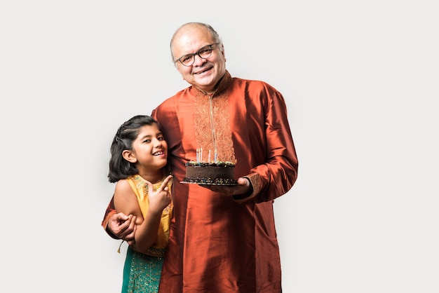 Indiase senior of oude man met kleindochter die verjaardag viert door kaarsen op taart te blazen terwijl hij etnische kleding draagt, geïsoleerd tegen een witte achtergrond