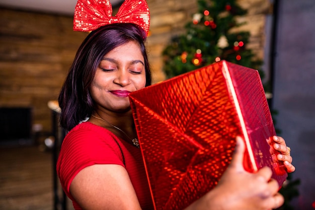 Indiase vrouw in rode jurk met grote doos met kom in sparren kerstboomverlichting in de woonkamer achtergrond