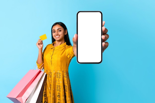 Indiase vrouw shopaholic houdt mobiele telefoon vast met een lege schermblauwe achtergrond