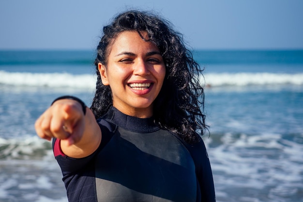 Indiase vrouwelijke surfer gekleed in een zwart nat pak staat in de buurt van de waterkant met één hand wijzend op de camera