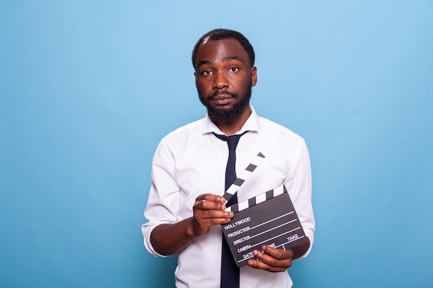 Indie african american movie producer with open clapperboard in hands looking confident. Independent successful filmmaker holding clapper standing proud over blue background.