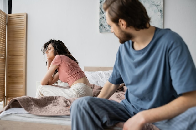 家のボーイフレンドの近くのベッドに座っているパジャマを着た無関心な女性
