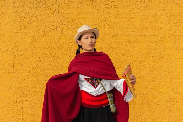 Indigenous woman with hat and red shawl holding a panpipe with rustic yellow wall