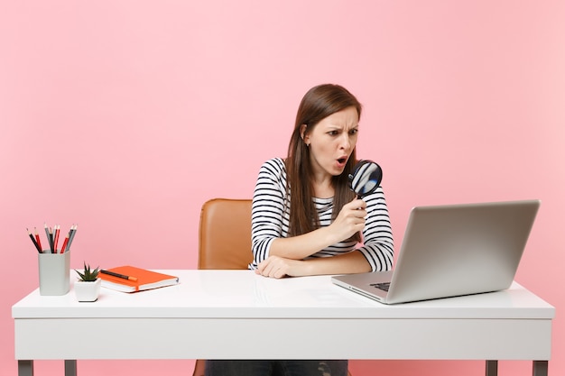 Indignant angry woman looking through magnifying glass on pc laptop while sitting and working on project at office isolated on pastel pink background. Achievement business career concept. Copy space.