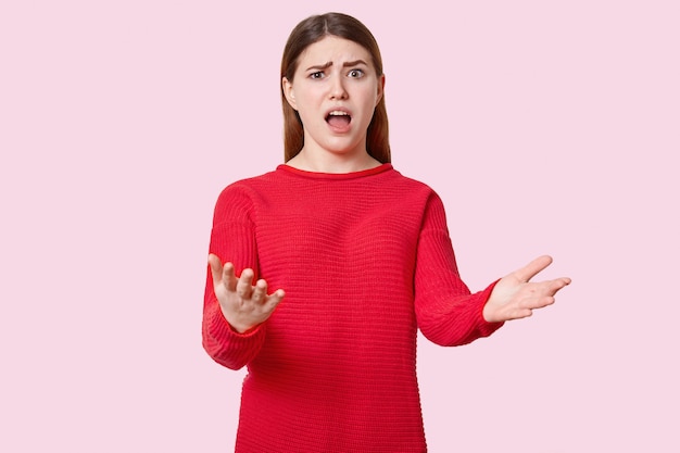 Indignant beautiful young woman gestures with puzzlement, opens mouth widely, raises eyebrows, dressed in red clothes, isolated on rosy wall. So what to do in this difficult situation?