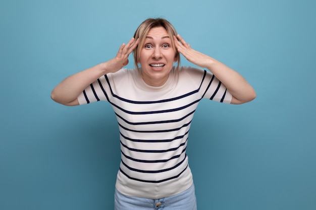 Indignant blond girl in a striped sweater is surprised on a blue background with copy space