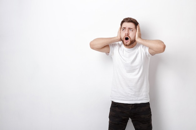 Indignant young man with dark hair and the beard in white t-shirt shouts in front of a white wall